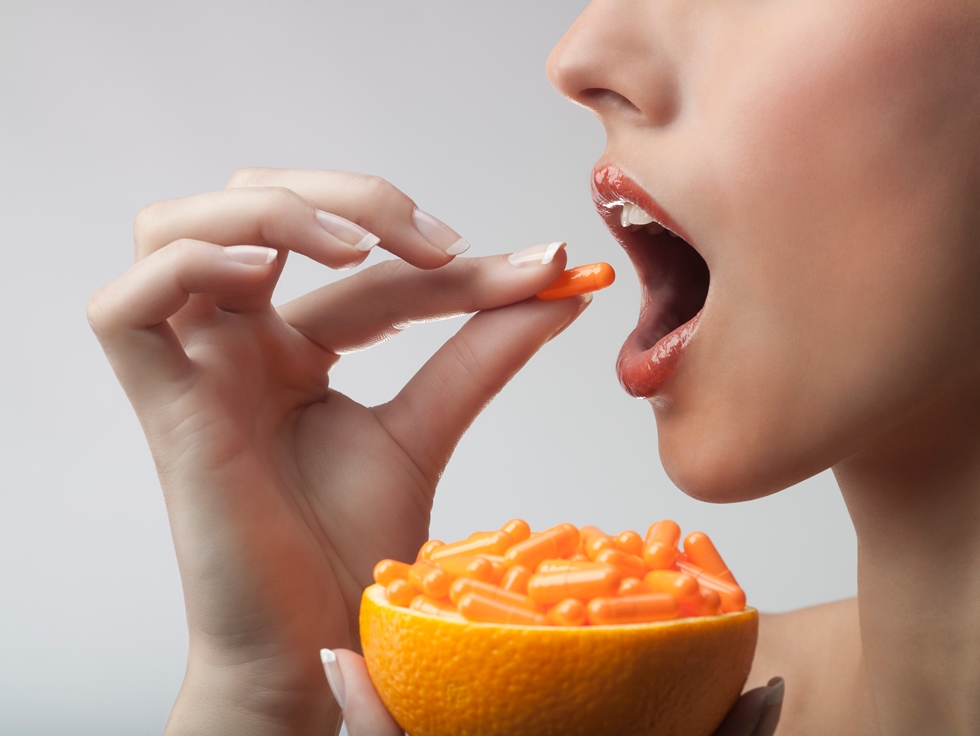 Žena stavlja kapsulu u usta dok su drugoj ruci drži pola naranče ispunjenom kapsulama.