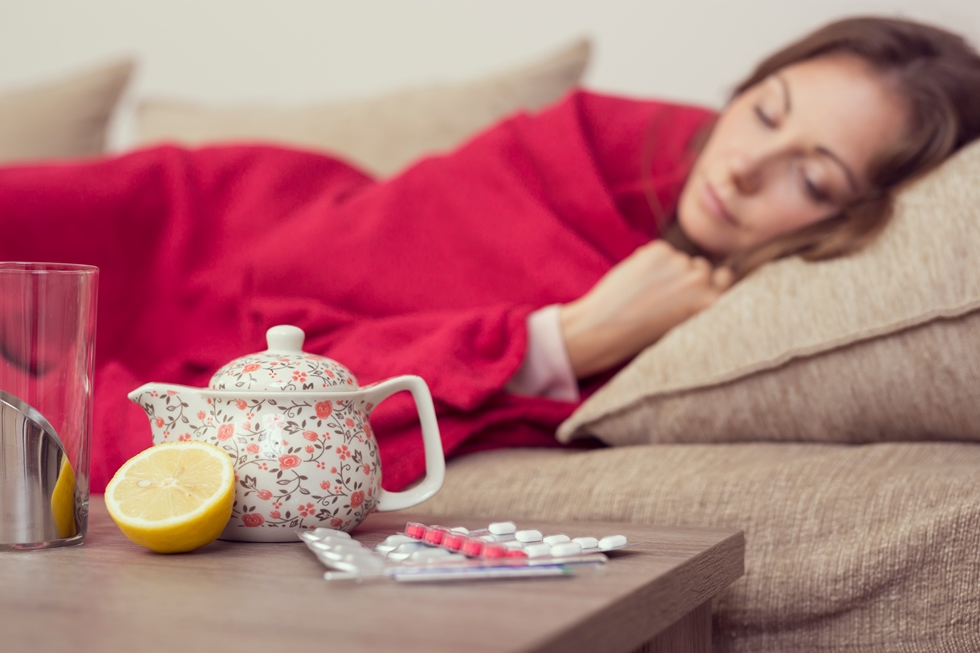 Žena pokrivine dekom spava na kauču, a na stoliću stoji čajnik, pola limuna i tablete.