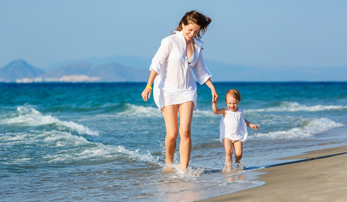 Mama i dijete hodaju po plićaku na pješćanoj plaži.