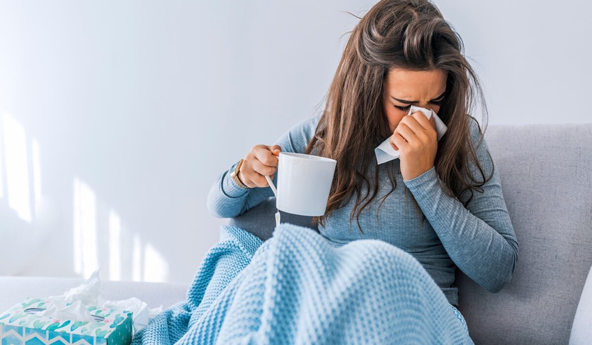 Žena puše nos u maramicu dok u drugoj ruci drži čaj dok sjedi na kauču pokrivena dekom.