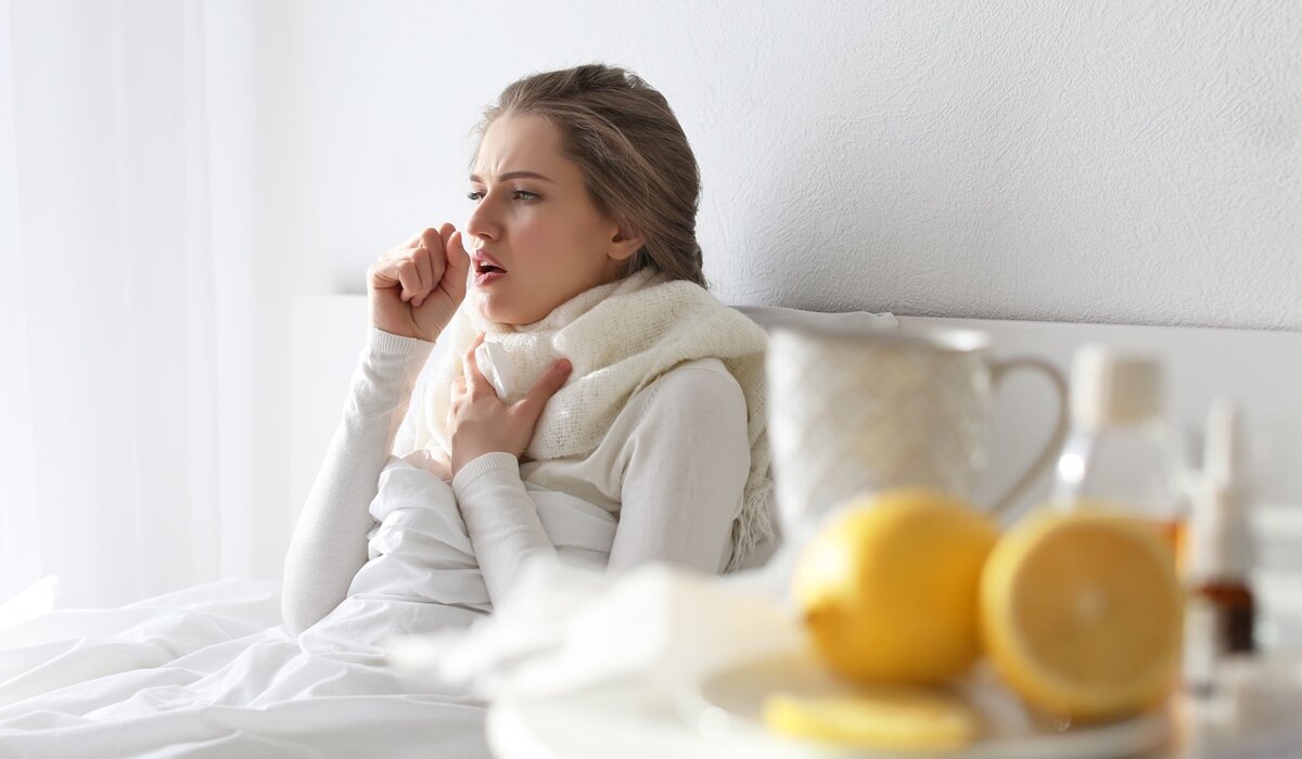 Žena u krevetu se drži za vrat dok kašlje i okružena je proizvodima protiv prehlade.